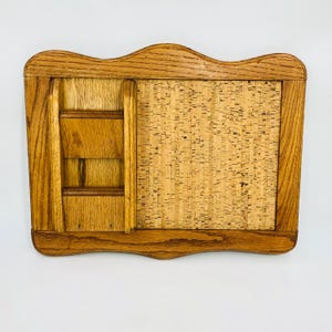 May include: A wooden wall-mounted organizer with a corkboard and small shelves. The frame is made of light brown wood with a wavy top edge. The corkboard is rectangular and takes up most of the space. The shelves are on the left side.