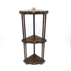 May include: A dark brown wooden corner shelf unit featuring three triangular tiers connected by vertical dowels. The top tier includes a small brass-colored metal ring centered at the back edge for mounting purposes, showcasing a smooth finish.