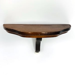 May include: A small, dark brown wooden shelf with a curved front edge and a single black support. The shelf has a groove cut into the top surface. The support is black with a white tip.