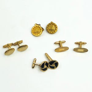 May include: A collection of gold-toned cufflinks in various designs. Styles include oval, circular, and those with black enamel accents. The cufflinks are displayed on a white surface, showcasing their different shapes and textures.