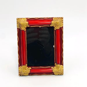 May include: A rectangular red picture frame with gold-colored floral corner accents. The frame has a glossy finish and a black interior, designed to hold a photograph or artwork. The frame is a decorative item.