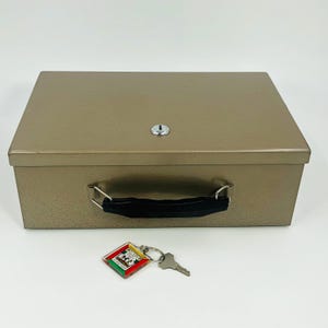 May include: A tan metal cash box with a black handle and a key lock. A key and a keychain with a colorful design are next to the box. The box is rectangular and has a simple, functional design.