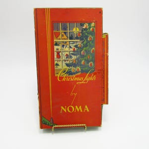 May include: Vintage Christmas lights box by NOMA. The red box features a window scene with a Christmas tree and the words "Christmas lights" in a decorative font. The box is displayed on a gold-colored wire stand.