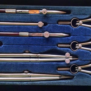 May include: A vintage drafting set in a black case with a blue velvet interior. The set includes several silver-toned compasses and dividers, along with drawing pens with brown and silver accents. The tools are neatly arranged.