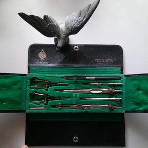 May include: An open black leather case with a green velvet interior, containing several drafting tools. A decorative bird statue sits above the case. The case has text that reads "Technical Supply Co. Scranton, Pa. Made in Germany."