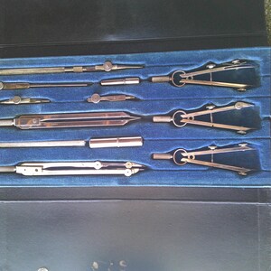 May include: A vintage drafting set in a black case with a blue velvet interior. The set includes several compasses, dividers, and other drawing tools made of metal. The case is open, revealing the tools neatly arranged.