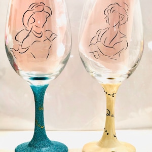 May include: Two wine glasses with a teal and cream glitter finish. The glasses are decorated with black line drawings of Aladdin and Jasmine. The teal glass has the words "To New" written on it in gold. The cream glass has the words "Don't Dance" written on it in black.