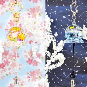 Puede incluir: Dos llaveros decorativos. Uno presenta un personaje de dibujos animados dentro de una campana transparente con una borla blanca. El otro llavero tiene una campana azul con una borla negra. Ambos tienen un cierre dorado en forma de flor o de luna.