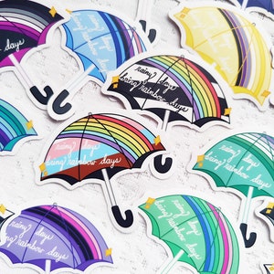 May include: A collection of colorful rainbow umbrella stickers with the text "rainy days bring rainbow days". The stickers are printed on white paper and have a glossy finish.