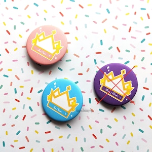 Puede incluir: Tres chapas circulares con un gráfico de corona amarilla. La chapa rosa muestra el texto she/her, la azul he/him y la morada they/them con una marca de X roja dibujada sobre toda la superficie de la chapa.