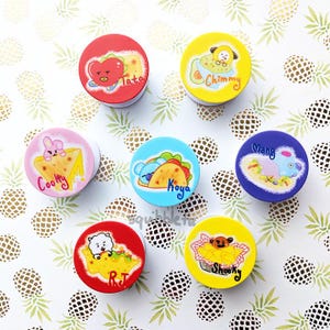 May include: Seven colorful phone grips with cartoon characters from the K-pop group BTS. Each grip features a different member of the group and a food-themed design. The characters include:  Cooky, Tata, Chimmy, Koya, RJ, Shooky, and Mang.