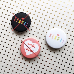 May include: Three circular pin-back buttons. One black button features the text SENPAI in rainbow colors with a red heart. One pink button features a white heart with the text hentai with senpai. One white button features the text SENPAI in rainbow colors with a red heart.