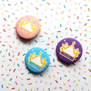 Op de afbeelding: Drie ronde pronoun-spelden in roze, blauw en paars. Elke speld heeft een gouden kroonontwerp met de voornaamwoorden "she/her", "he/him" en "they/them" eronder. De achtergrond is wit met kleurrijke confetti.