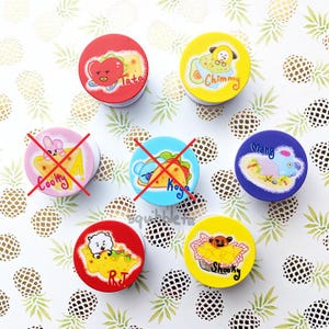 Pode incluir: Sete objetos circulares com personagens de desenhos animados e ilustrações de comida. Dois objetos estão marcados com um X vermelho. Os designs incluem Tata, Chimmy, Cooky, Koya, Mang, RJ e Shooky, rotulados com seus nomes em escrita colorida.