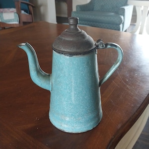 May include: A vintage, light blue enamelware coffee pot with a dark brown lid and handle. The pot has a spout and a curved handle. The enamel has a speckled appearance, and the pot appears to be well-used.