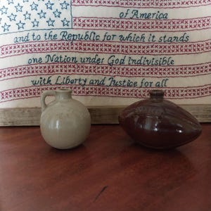 May include: Two small, ceramic jugs sit on a wooden surface. The jug on the left is light beige with a handle. The jug on the right is dark brown. Behind them is a framed textile with the text "of America" and "and to the Republic for which it stands."