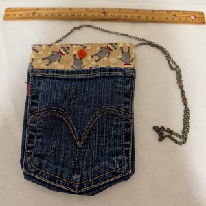 May include: A denim pocket purse with a chain strap. The purse is made from a repurposed blue jean pocket, with a sock monkey print fabric lining the top. A wooden ruler is placed above the purse.