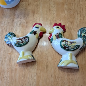 May include: Two ceramic rooster figurines featuring a cream-colored base with hand-painted details in red, yellow, and green. Each rooster has a prominent red comb, a yellow beak, and stylized green and brown wing feathers on a white body.