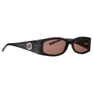 Puede incluir: Gafas de sol marrones oscuras con montura rectangular y lentes marrones. Las gafas de sol presentan un emblema plateado en el lateral de la montura. Un accesorio elegante.