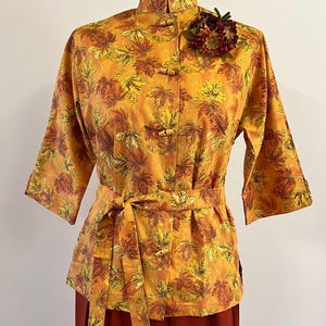 May include: A yellow and orange blouse with a leaf print and a brown belt. The blouse has a mandarin collar and wooden buttons. A red and brown flower pin is on the collar.