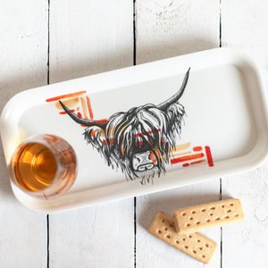 May include: White ceramic tray with a black and white illustration of a Highland cow's head. The tray has a red and orange abstract design. A glass of amber liquid and two shortbread cookies are on the tray.
