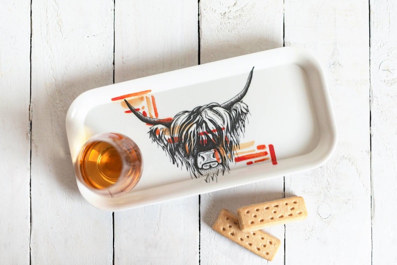 May include: White ceramic tray with a black and white illustration of a Highland cow's head. The tray has a red and orange abstract design. A glass of amber liquid and two shortbread cookies are on the tray.