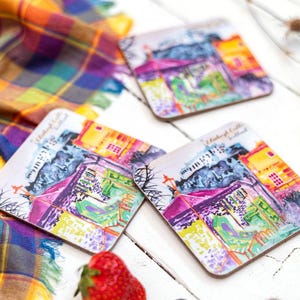 May include: A set of square coasters featuring watercolour illustrations of Edinburgh Castle, Scotland. The coasters are brightly coloured and are arranged on a white wooden surface with a plaid scarf, a strawberry, and a lemon slice.