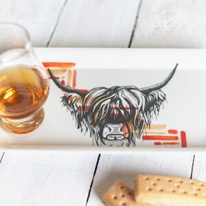 May include: A white ceramic tray with a black and white illustration of a Highland cow with orange and red abstract designs. A glass of amber liquid and two shortbread cookies are on the tray.