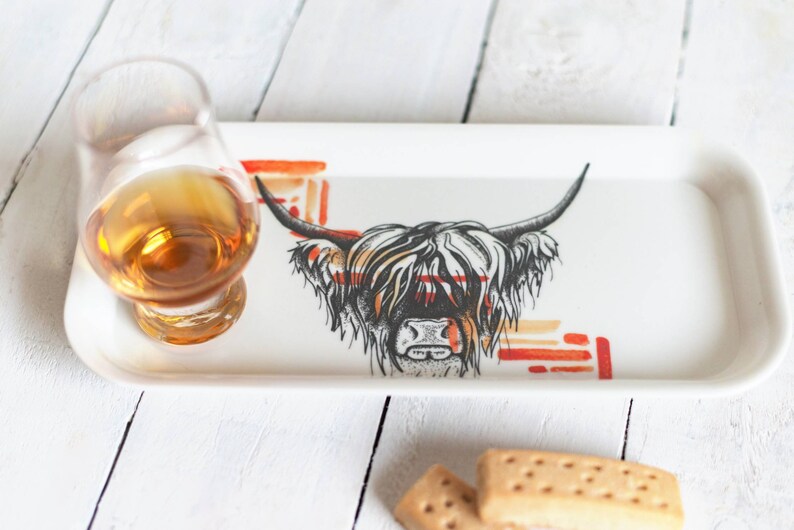 May include: A white ceramic tray with a black and white illustration of a Highland cow with orange and red abstract designs. A glass of amber liquid and two shortbread cookies are on the tray.