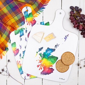 May include: Two white wooden chopping boards with a rainbow watercolour design of the outline of Scotland. The boards have a handle and the word "Scotland" is printed on the bottom of one board.