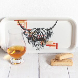 May include: A white ceramic tray with a black and white illustration of a Highland cow's head. The cow's fur is white with black accents. The tray has a red and orange geometric pattern in the background. A glass of amber liquid and two shortbread cookies are on a white surface in front of the tray.