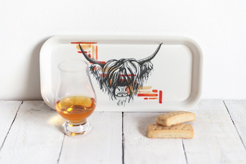 May include: A white ceramic tray with a black and white illustration of a Highland cow's head. The cow's fur is white with black accents. The tray has a red and orange geometric pattern in the background. A glass of amber liquid and two shortbread cookies are on a white surface in front of the tray.