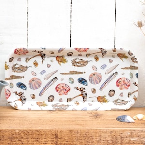 May include: A rectangular serving tray featuring a white background with watercolour illustrations of seashells, sea urchins, and seaweed. The tray is on a wooden surface, next to a blue teapot and a vase with dried flowers.