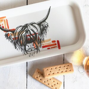 May include: A white ceramic tray with a black and white illustration of a Highland cow's head. The cow's fur is depicted in a stylized pattern with red and orange accents. The tray is on a white wooden surface with two shortbread cookies and a glass of amber liquid.