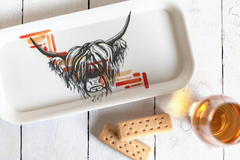 May include: A white ceramic tray with a black and white illustration of a Highland cow's head. The cow's fur is depicted in a stylized pattern with red and orange accents. The tray is on a white wooden surface with two shortbread cookies and a glass of amber liquid.