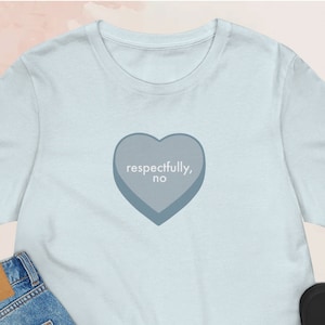May include: Light blue t-shirt with a gray heart graphic that says "respectfully, no". The t-shirt is paired with blue jeans, a beige beanie, and black sneakers.