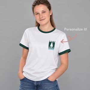 Peut inclure: Un t-shirt blanc à bordures vert foncé sur le col et les manches. La poitrine présente un motif carré vert foncé avec une icône de sapin blanc au-dessus du texte "CAMP WALDEN" et des lignes ondulées bleues. Le texte "Personalize it!" est visible.