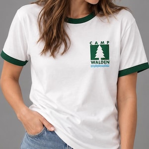 Può includere: Una t-shirt bianca con bordi verde scuro sul colletto e sulle maniche. Sul petto è presente una grafica con un quadrato verde contenente l'icona di un pino bianco, la scritta CAMP WALDEN in nero e linee ondulate blu sottostanti.