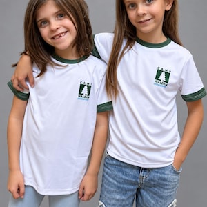 May include: A white short-sleeve ringer t-shirt with dark green trim on the collar and sleeve cuffs. The chest features a graphic logo with a green pine tree, the text CAMP WALDEN, and a blue wavy line underneath the tree.