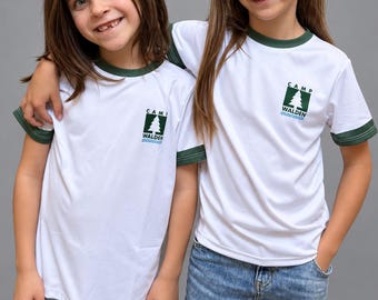 May include: A white short-sleeve ringer t-shirt with dark green trim on the collar and sleeve cuffs. The chest features a graphic logo with a green pine tree, the text CAMP WALDEN, and a blue wavy line underneath the tree.