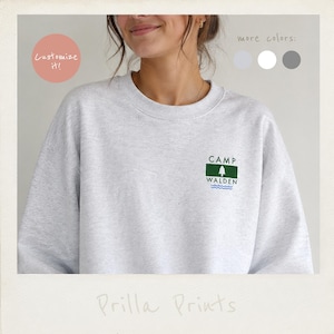 May include: A light gray crewneck sweatshirt featuring a small graphic on the left chest. The graphic includes a green rectangle with a white pine tree icon, the text "CAMP WALDEN" in black, and three blue wavy lines underneath the text.