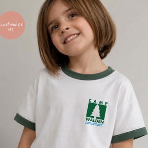 May include: A white ringer t-shirt with dark green trim on the collar and sleeve cuffs. The chest features a green square graphic containing a white pine tree illustration, the text CAMP WALDEN in green, and blue wavy lines at the bottom.