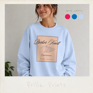 Può includere: Una felpa girocollo azzurro chiaro con una grafica rettangolare color pesca sul petto. La grafica riporta il testo Parker Knoll, 1993, Chardonnay e Napa Valley, insieme a un disegno al tratto che raffigura un paesaggio di vigneti.