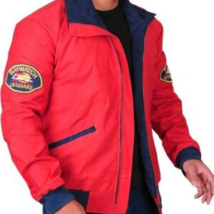 May include: A red jacket with a navy blue collar, cuffs, and interior lining. The jacket features a "Baywatch Lifeguard" patch on the left sleeve and a small patch on the right sleeve. The jacket has a zip closure and a navy blue pocket detail.