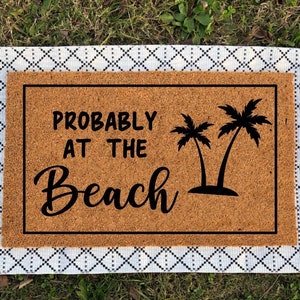 May include: A brown coir doormat with the text "Probably at the Beach" and two black palm trees. The doormat has white fringe around the edges.