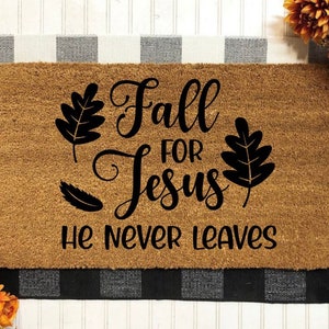 May include: A brown doormat with the text "Fall for Jesus He Never Leaves" in black lettering. The text is surrounded by black leaf designs.