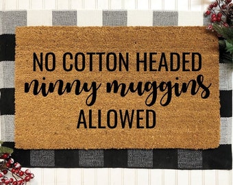 May include: A rectangular doormat made of natural coir fiber, featuring the phrase "NO COTTON HEADED ninny muggins ALLOWED" in black script. The mat is set on a black and white checkered rug, with holiday greenery in the corners.