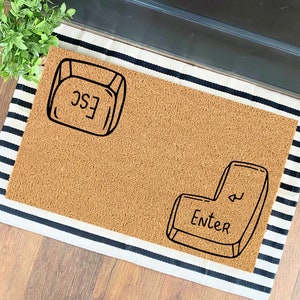 May include: A brown doormat with black and white stripes around the edges. The doormat has two black and white drawings of computer keys. One key says "ESC" and the other says "Enter".