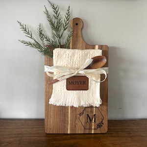 May include: A wooden cutting board with a handle, featuring a cream-colored towel, wooden spoon, and a leather tag that reads "MOYER". The board is decorated with a floral design and the letter "M". A sprig of greenery is tucked behind the towel.