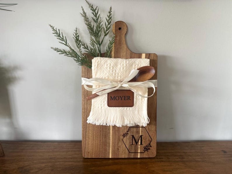 May include: A wooden cutting board with a handle, featuring a cream-colored towel, wooden spoon, and a leather tag that reads "MOYER". The board is decorated with a floral design and the letter "M". A sprig of greenery is tucked behind the towel.